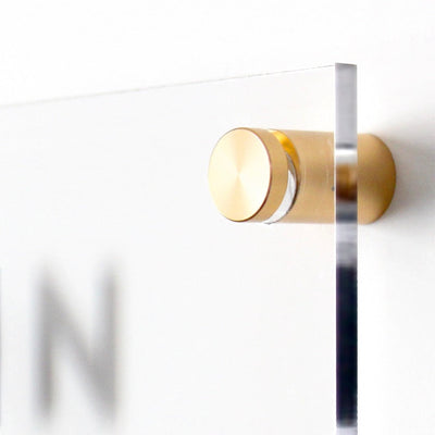 A close-up image features a Girl Friday acrylic Goal Chart | Sales Tracker for Real Estate mounted on a wall with gold fittings. The polished cylindrical hardware gleams, and a blurred letter N evokes customizable houses and innovative design ideas.