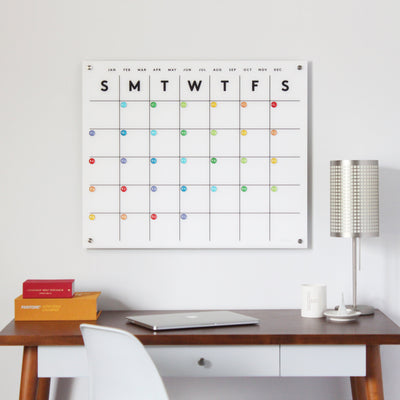 A minimalist desk holds a laptop and books beneath a Girl Friday Shops Magnetic Acrylic Monthly Calendar, featuring weekly planning grids and colorful dots under weekday initials. A modern lamp is on the desk, with a wet-erase marker nearby for updates.