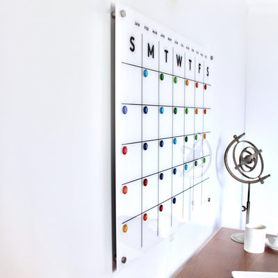 A Girl Friday Shop Magnetic Acrylic Monthly Calendar with colorful marker magnets marks weekdays. Beside it, a sleek metallic armillary sphere rests on a wooden desk against a pristine white wall.