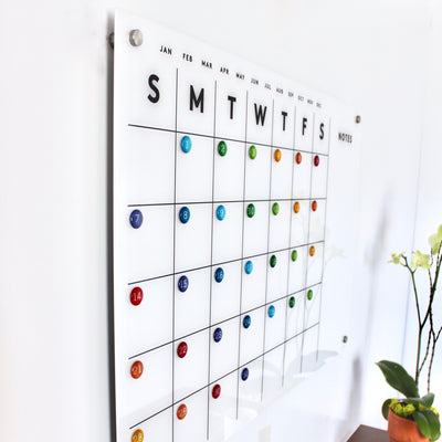 The Magnetic Acrylic Calendar with Side Notes by Girl Friday Shop features a monthly layout with colored magnets for each day and columns from Sunday to Saturday. Its premium dry erase surface complements nearby decor, while a small note section adds functionality.