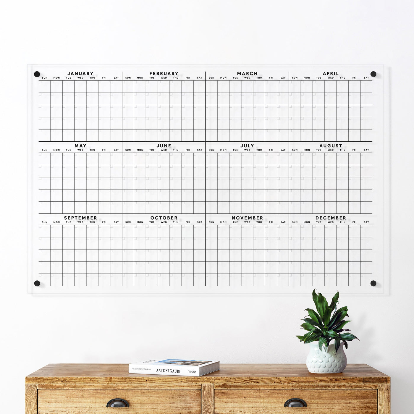The Clear Acrylic Yearly Wall Calendar | Horizontal by Girl Friday features a grid displaying all months from January to December. It looks perfect above a light wooden console table, pairing well with dry-erase markers for notes, alongside a potted plant and a book.