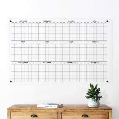The Clear Acrylic Yearly Wall Calendar | Horizontal by Girl Friday features a grid displaying all months from January to December. It looks perfect above a light wooden console table, pairing well with dry-erase markers for notes, alongside a potted plant and a book.