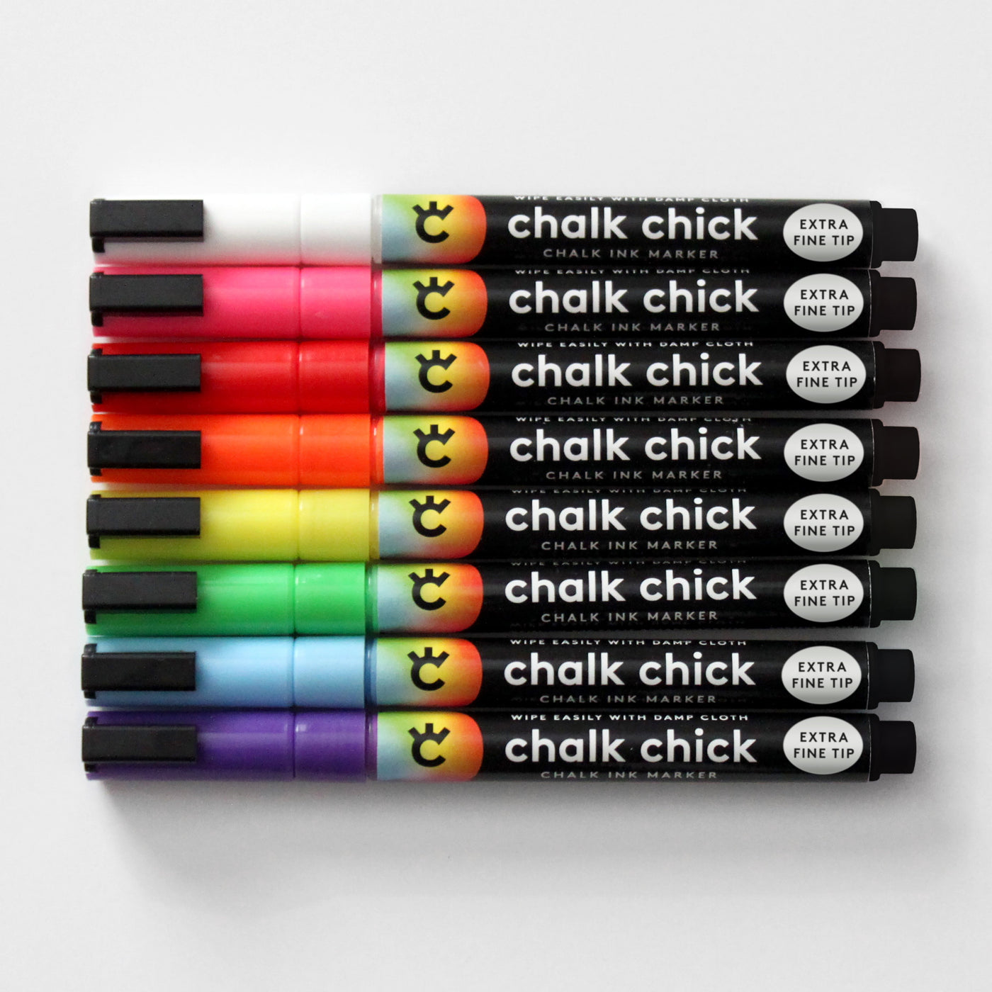 Eight vibrant Chalk Chick markers are lined up horizontally, featuring black, white, red, orange, yellow, green, blue, and purple. These wet-erase markers have black caps labeled Extra Fine Tip, perfect for the Clear Acrylic Monthly Fridge Calendar by Girl Friday Shop.