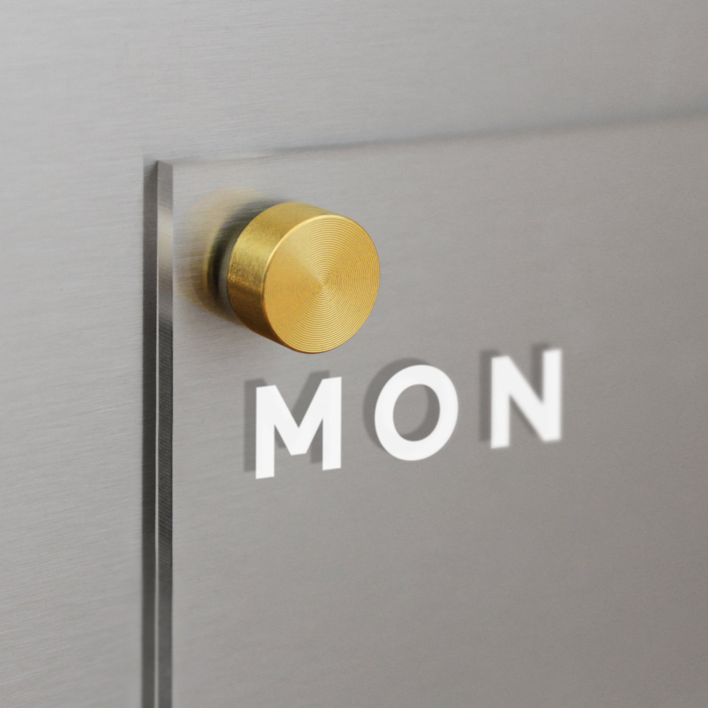 A close-up of a frosted glass panel etched with MON on the Acrylic Fridge Calendar by Girl Friday Shop, fastened with a round gold metal piece to a metallic surface.