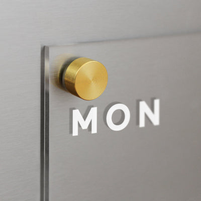 A close-up of a frosted glass panel etched with MON on the Acrylic Fridge Calendar by Girl Friday Shop, fastened with a round gold metal piece to a metallic surface.