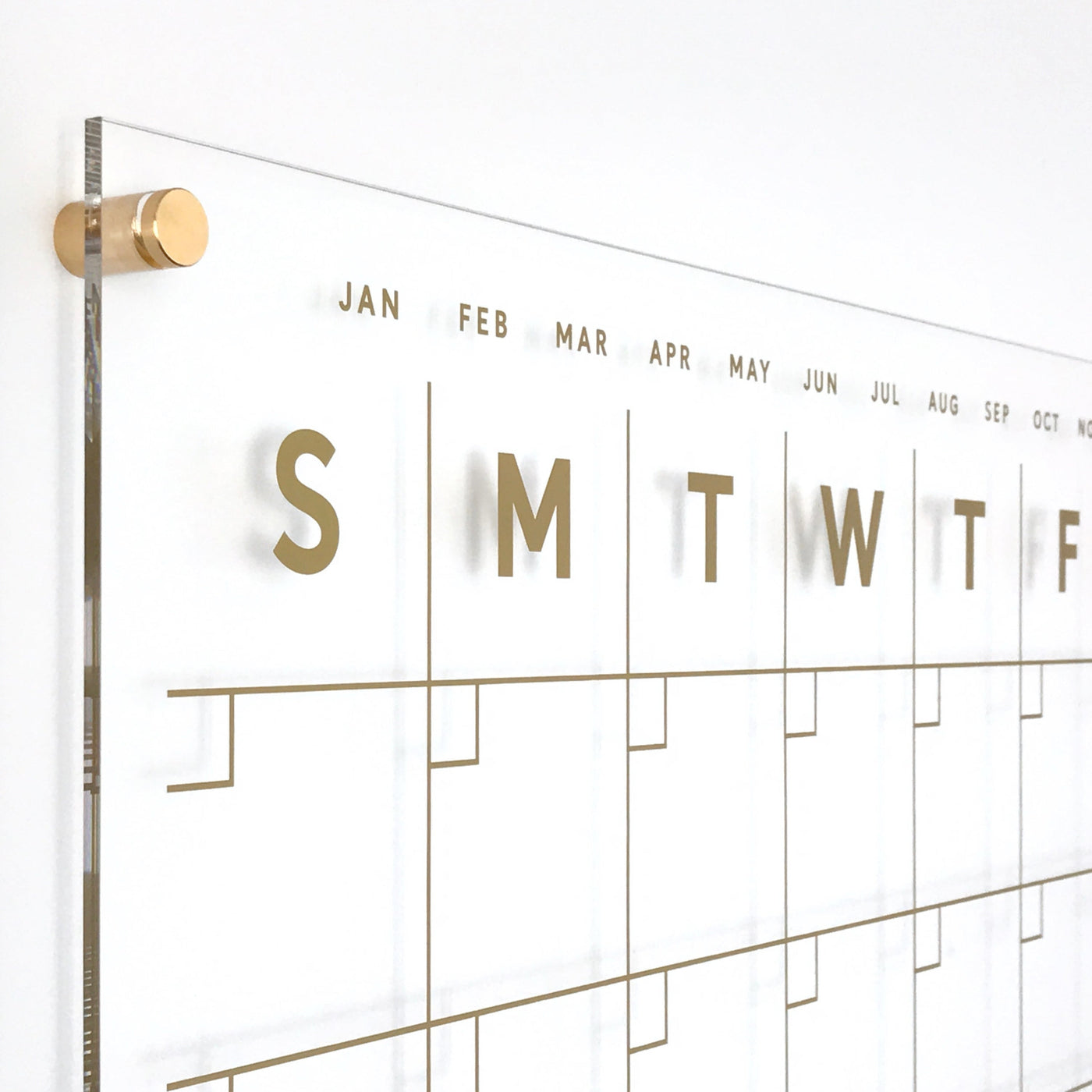 The Clear Acrylic Monthly Wall Calendar with Side Notes by Girl Friday Shop features polished edges and gold text for the days (Sunday to Saturday) and months. Gold standoff screws ensure secure wall mounting, making it ideal for wet-erase markers.