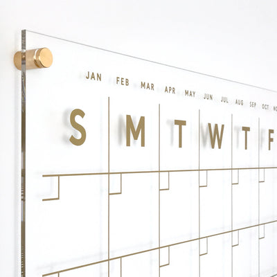 The Clear Acrylic Monthly Wall Calendar with Side Notes by Girl Friday Shop features polished edges and gold text for the days (Sunday to Saturday) and months. Gold standoff screws ensure secure wall mounting, making it ideal for wet-erase markers.