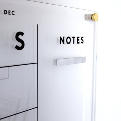 Close-up of a transparent acrylic wall calendar with a minimalistic design using neodymium cube magnets and metallic mounting hardware. The NOTES section, enhanced by Girl Fridays metal strips for magnets, blends functionality with modern elegance.