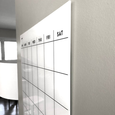 A close-up of the White Acrylic Monthly Calendar by Girl Friday Shop shows days labeled: Sun, Mon, Tue, Wed, Thu, Fri, and Sat. The glossy dry erase surface is mounted on a light wall in a bright room with a white chair visible. Ideal for non-magnetic fridges.