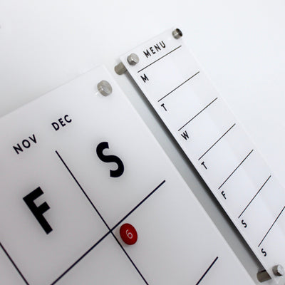 A calendar shows late November and early December, marked on the 6th. Beside it is Girl Friday Shops sleek Magnetic Acrylic Menu Board, featuring abbreviated weekdays for weekly planning. Both items are white with black text.