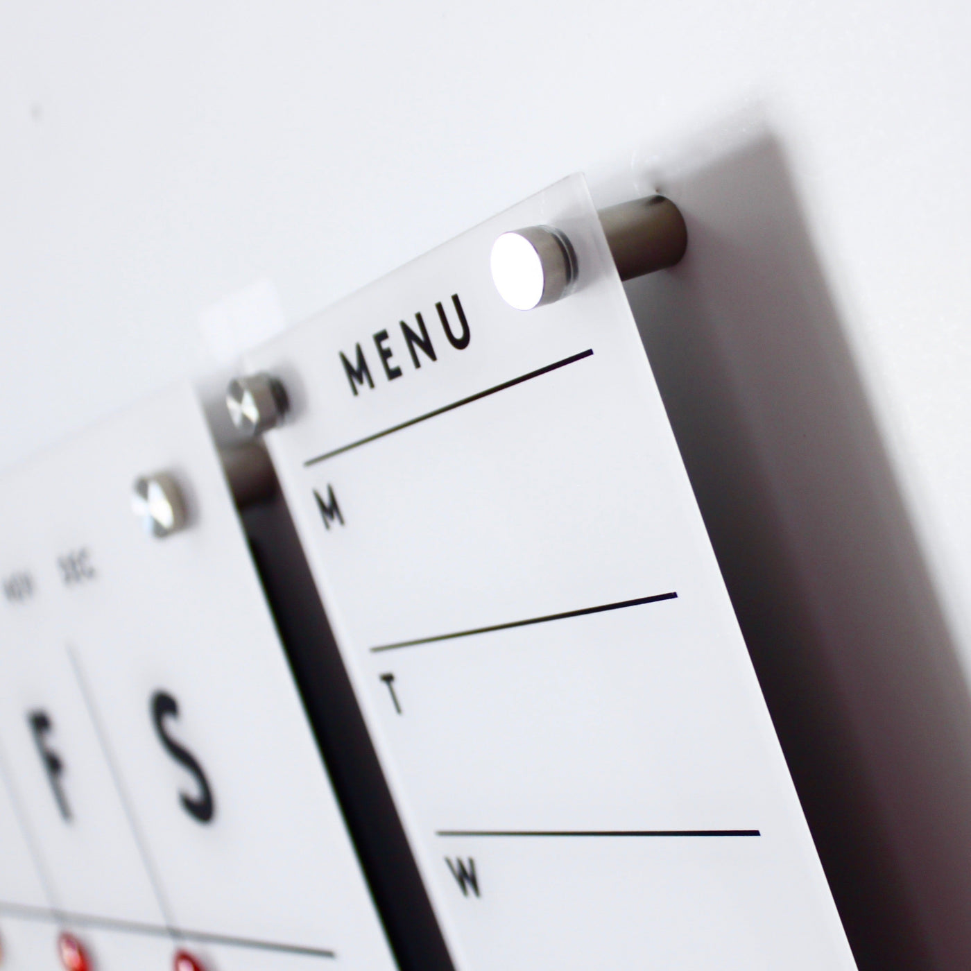 Close-up of the Girl Friday Shop Magnetic Acrylic Menu Board with sections for Monday to Wednesday. MENU is prominently displayed at the top, and its mounted on a white wall using metal pegs.