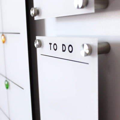 Close-up of a Girl Friday Shop Magnetic Acrylic Menu Board with a clean slate for tasks, affixed to the wall using metallic knobs, showcasing its sleek functionality.