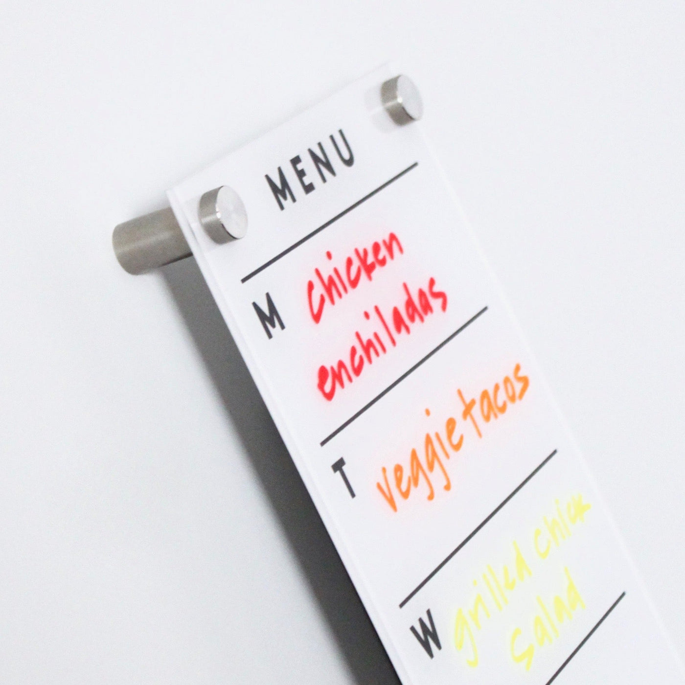 The Girl Friday Shops Magnetic Acrylic Menu Board, mounted with stylish silver pegs, lists three days of meals in vibrant hues: Monday has red chicken enchiladas, Tuesday features orange veggie tacos, and Wednesday offers a yellow grilled chicken salad.