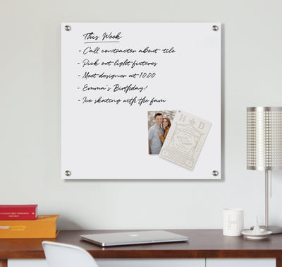 A Magnetic Acrylic Memo Board from Girl Friday Shop lists tasks, like calling a contractor and Emma’s birthday, in black handwriting. A small photo and card are attached. Nearby, a metal lamp and stack of books sit on the desk.