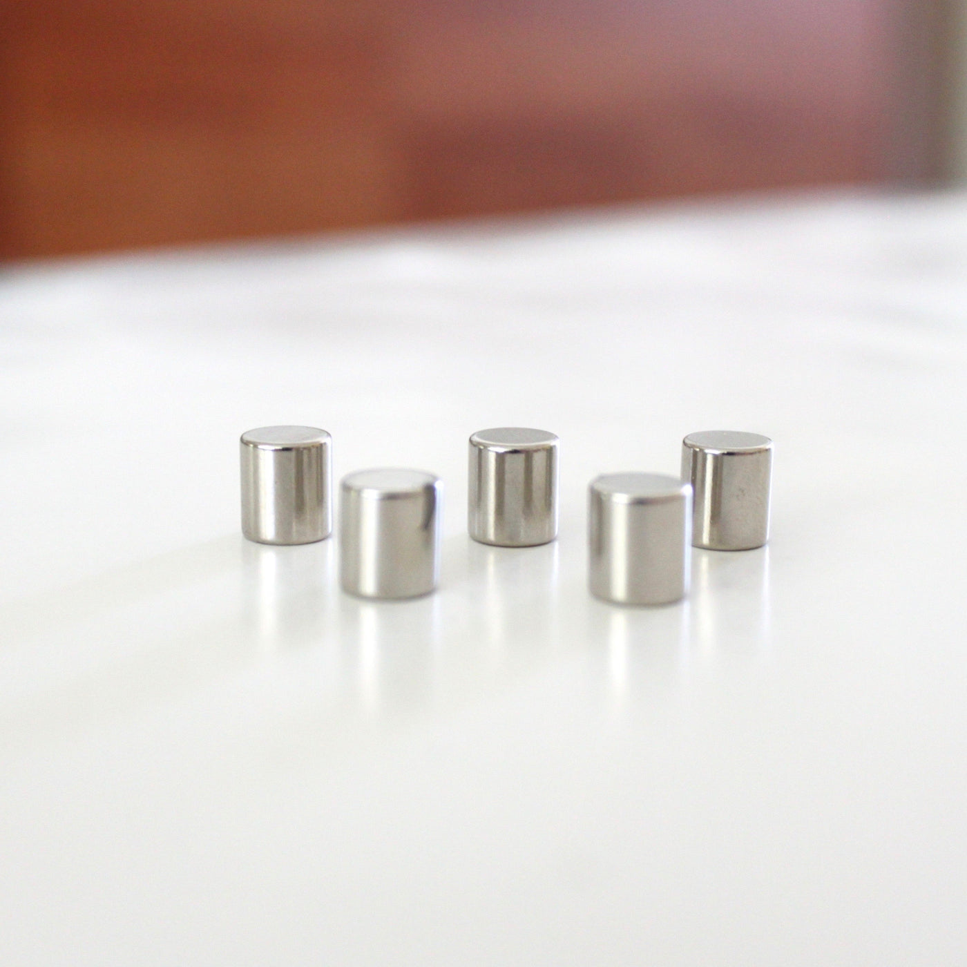 Five metallic Cylinder Magnets from Girl Fridays Magnetic Surface Upgrade for Framed Boards lie on a white surface, their strength clear against a warm-toned blur, ideal for securing items.
