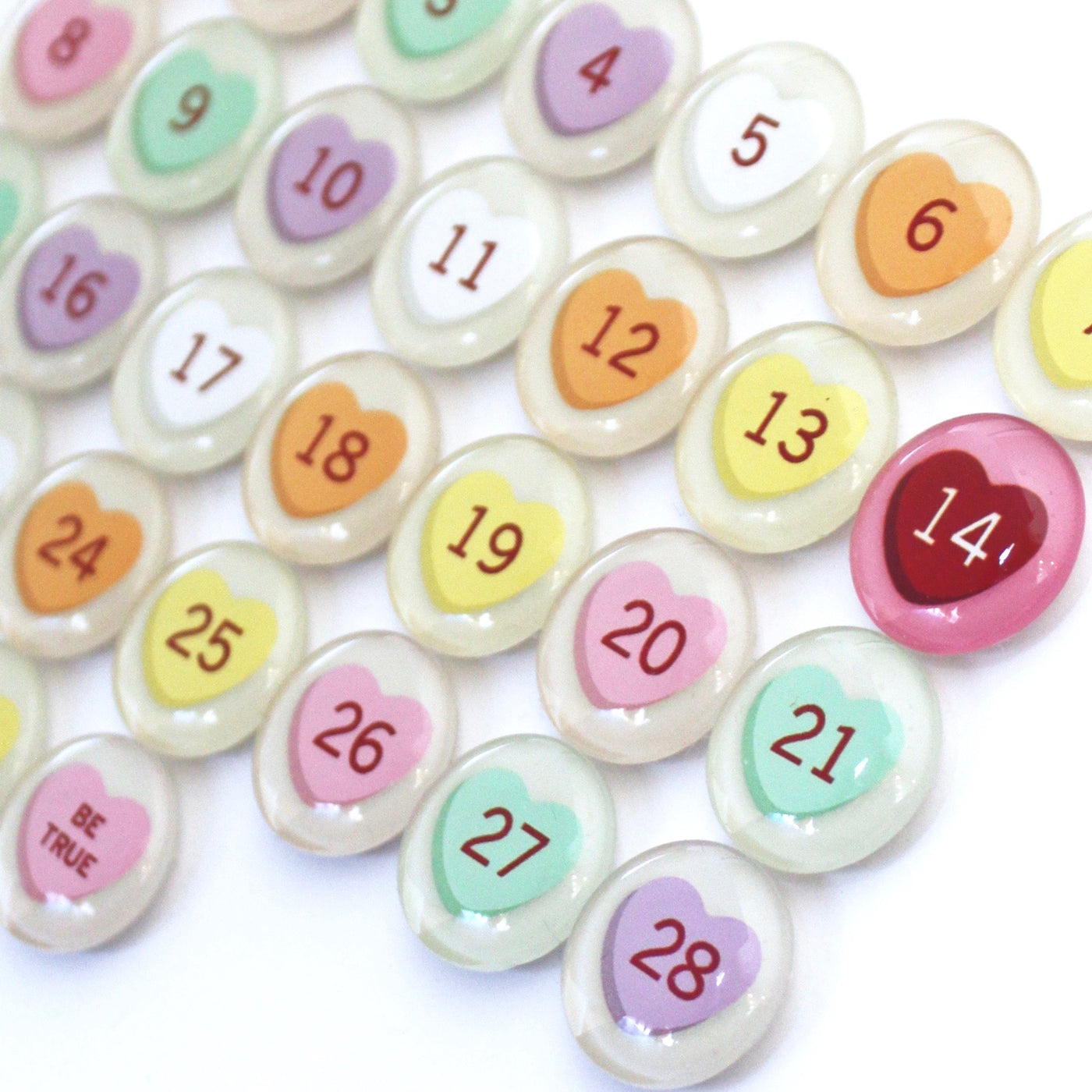 Girl Fridays Number Magnets for February feature colorful, pastel, heart-shaped magnets numbered 1 to 28 on a white surface. The number 14 is highlighted in red, and BE TRUE stands out among these Valentines Day conversation hearts.