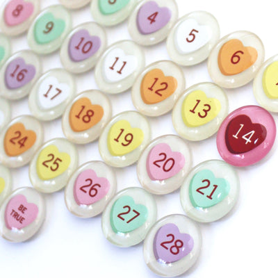 Girl Fridays Number Magnets for February feature colorful, pastel, heart-shaped magnets numbered 1 to 28 on a white surface. The number 14 is highlighted in red, and BE TRUE stands out among these Valentines Day conversation hearts.