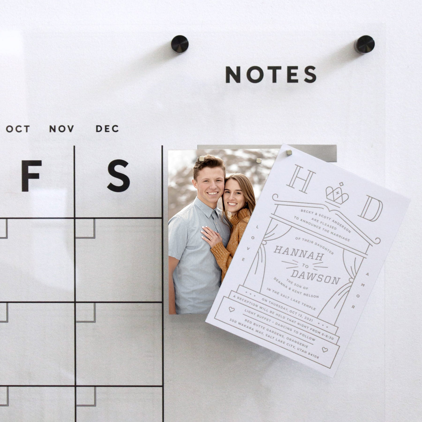 A Girl Friday acrylic wall display features an overlapping engagement photo of a smiling couple, secured with sleek neodymium cube magnets held by Metal Strips, and a wedding invitation card with elegant typography and floral design.
