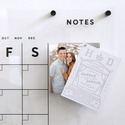 A Girl Friday acrylic wall display features an overlapping engagement photo of a smiling couple, secured with sleek neodymium cube magnets held by Metal Strips, and a wedding invitation card with elegant typography and floral design.