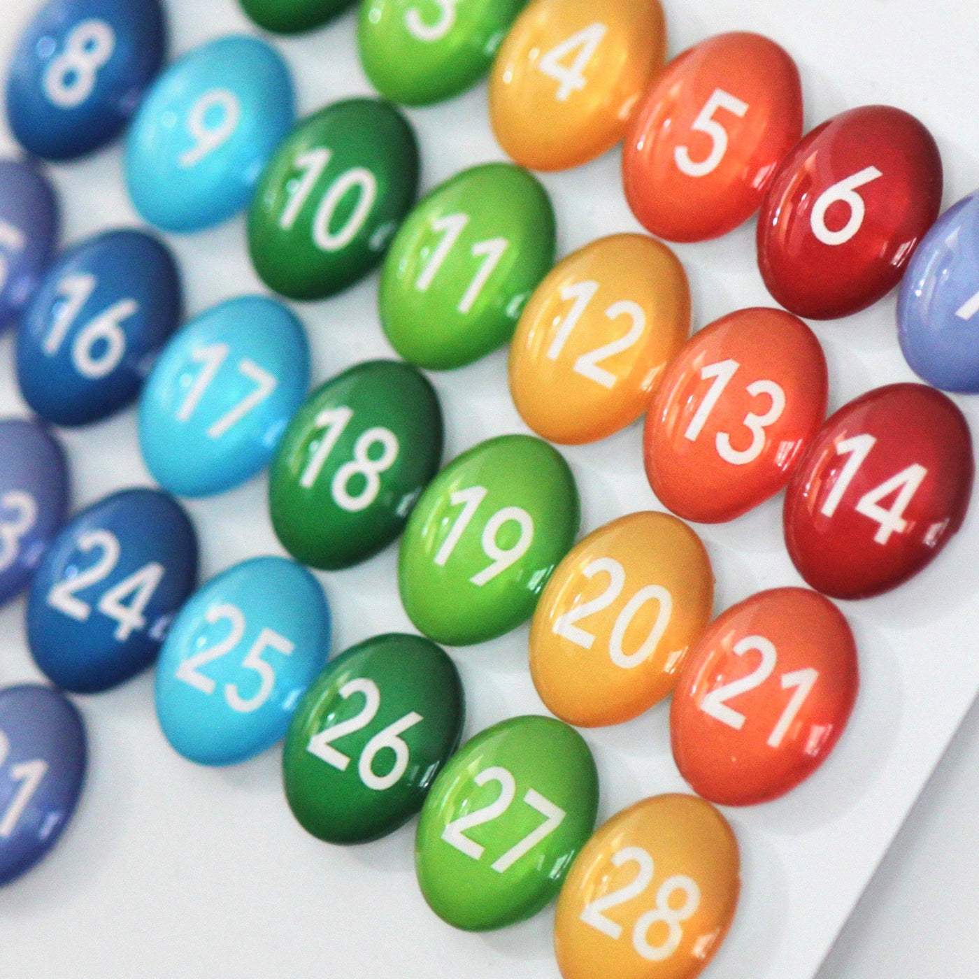 Girl Fridays Number Magnets - Rainbow, numbered 1 to 28, are colorful circular glass magnets arranged in a grid. They transition through blue, green, yellow, orange, and red shades to create a vibrant gradient effect similar to rainbow glass magnets.