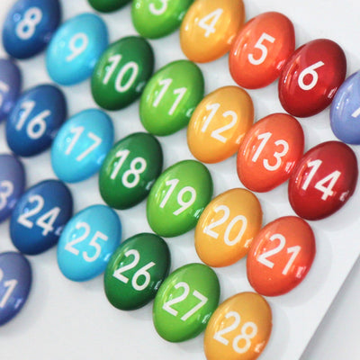 Girl Fridays Number Magnets - Rainbow, numbered 1 to 28, are colorful circular glass magnets arranged in a grid. They transition through blue, green, yellow, orange, and red shades to create a vibrant gradient effect similar to rainbow glass magnets.