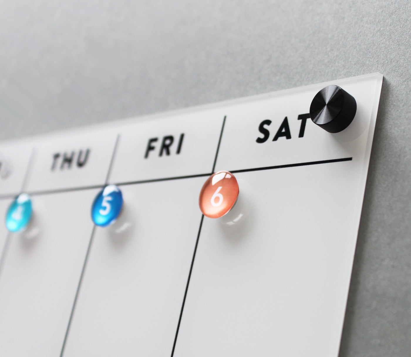 The White Acrylic Fridge Calendar by Girl Friday Shop features a dry-erase calendar board. Colorful magnets mark days: blue on Thursday and Friday, orange on Saturday. It resembles an acrylic fridge calendar with a black knob at the top right corner.