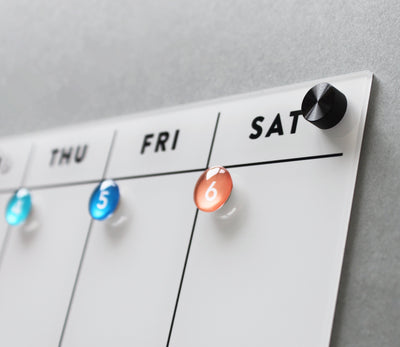 The White Acrylic Fridge Calendar by Girl Friday Shop features a dry-erase calendar board. Colorful magnets mark days: blue on Thursday and Friday, orange on Saturday. It resembles an acrylic fridge calendar with a black knob at the top right corner.