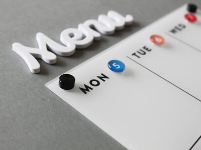 A close-up of the White Acrylic Weekly Fridge Menu Board with Notes by Girl Friday Shop shows Menu in raised white letters. It features sections for Mon to Wed, marked with colorful round magnets, ideal for a magnetic fridge or dry erase surface.