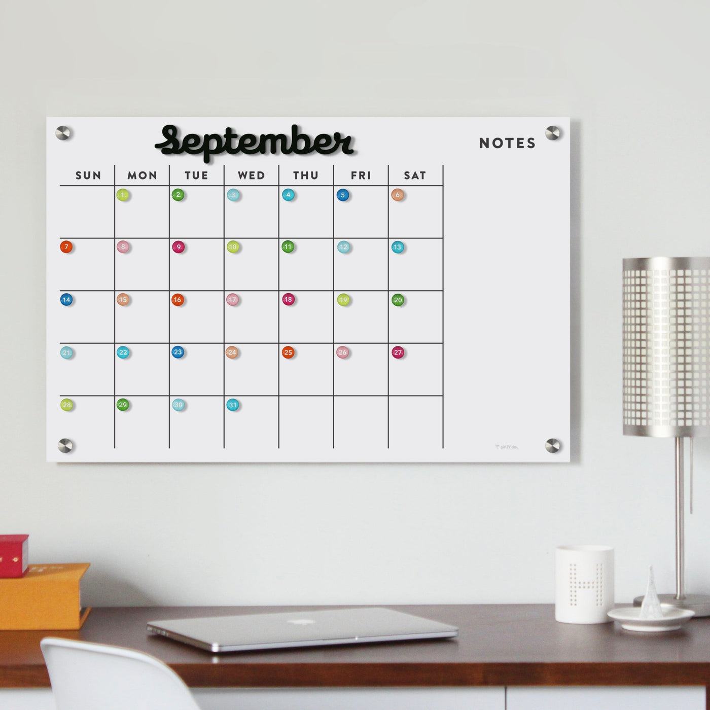 The Girl Friday Shops Magnetic Acrylic Calendar includes black magnetic months and a dry-erase surface. Perfect for September, it features colorful markers on dates and a Notes section, seamlessly blending with your light-colored wall beside a lamp and your book container for a modern desk setup.