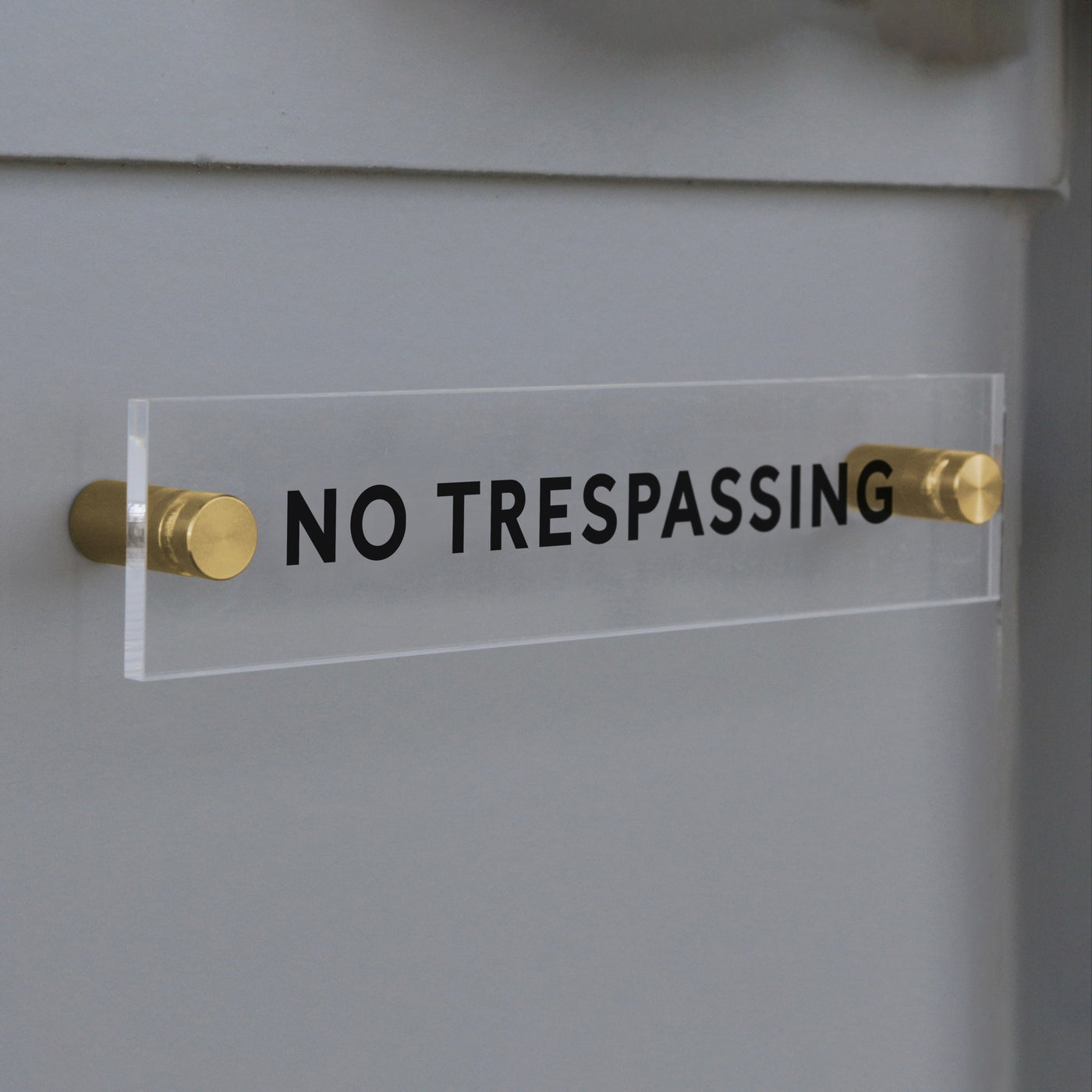 The No Trespassing Acrylic Sign by Girl Friday Shop displays bold black text and is elegantly mounted on a gray wall with two brass standoffs.