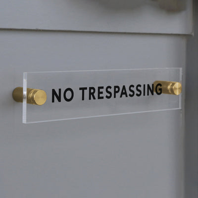 The No Trespassing Acrylic Sign by Girl Friday Shop displays bold black text and is elegantly mounted on a gray wall with two brass standoffs.