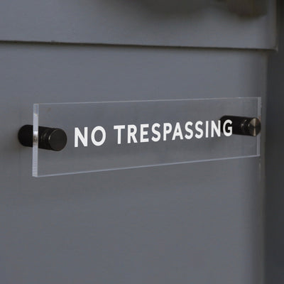A Girl Friday Shops No Trespassing Acrylic sign is mounted on a gray wall using two black metal standoffs.