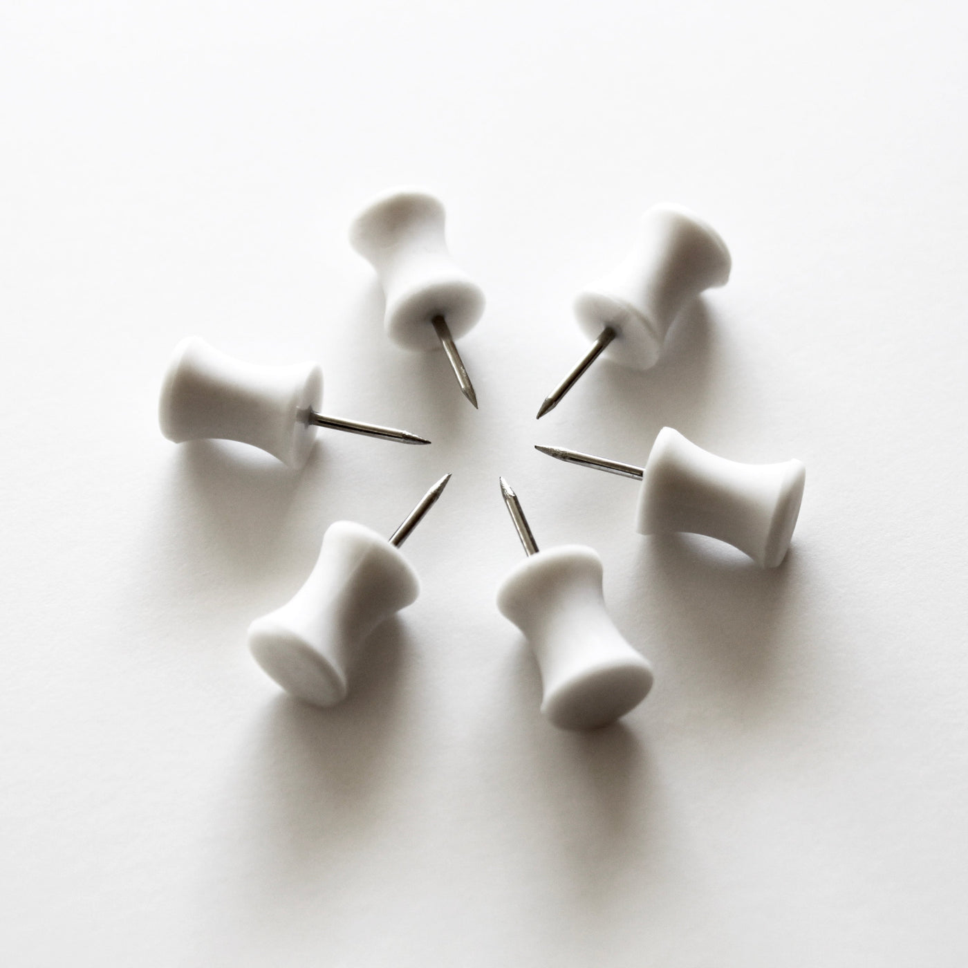 Six modern push pins by Girl Friday, with sleek rubber grips, are elegantly arranged in an hourglass shape on a pristine white surface.