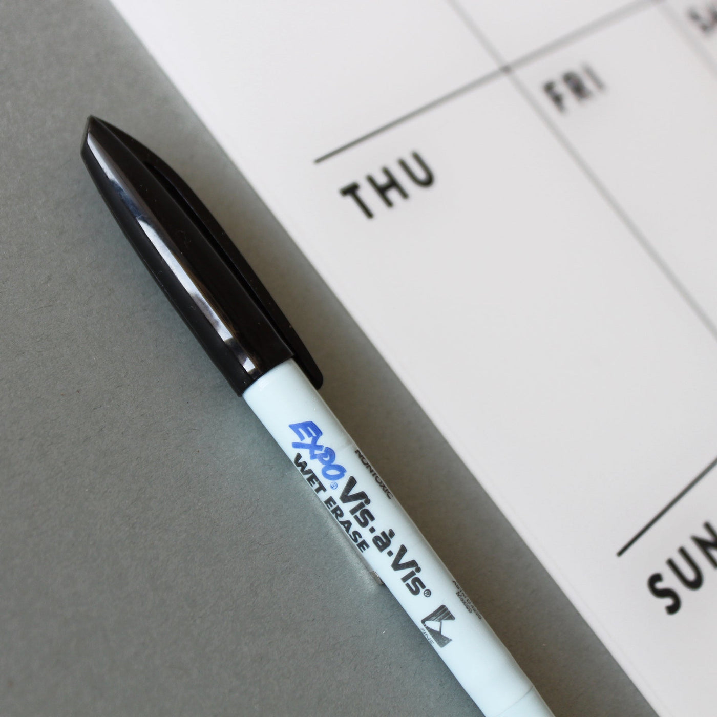 A white Expo Vis-à-Vis wet erase marker with a black cap rests on a gray surface next to a sleek Girl Friday Shop Chore Chart for 1 or 2 children, showing Thursday to Sunday.