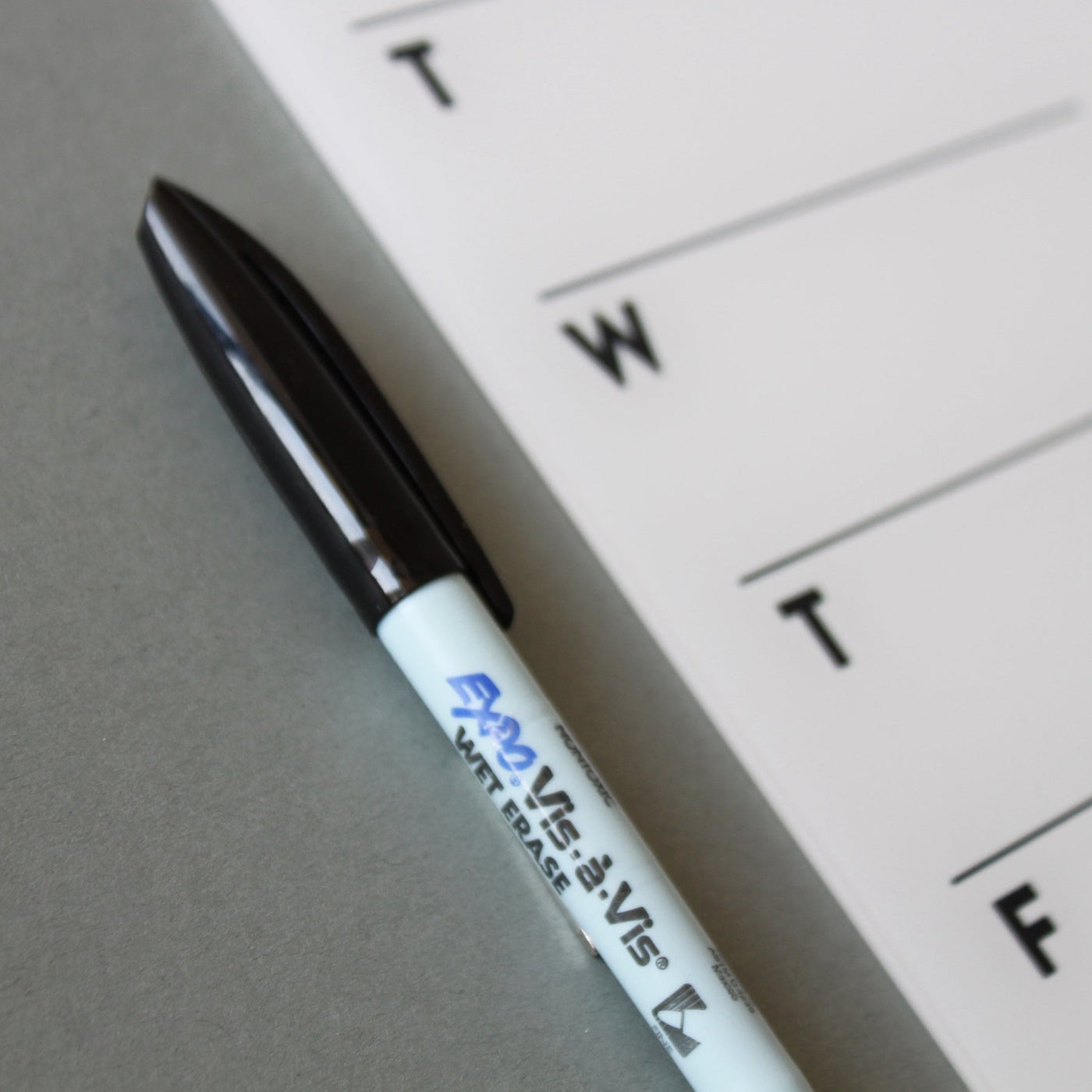 A black Expo Vis-a-Vis Wet Erase marker pen lies diagonally on a gray surface beside a white paper calendar, suggesting its use on the Girl Friday Shops White Acrylic Weekly Fridge Menu Board with visible letters T and W.