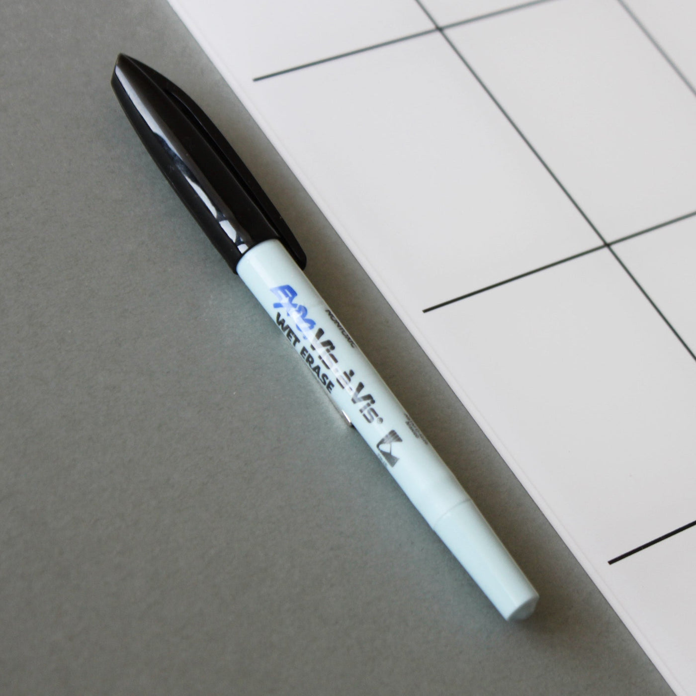 Beside a Girl Friday Shop White Acrylic Fridge Calendar with bottom notes, a black-capped whiteboard marker featuring blue lettering lies on a gray surface, adjacent to the edge of a grid-patterned whiteboard, ideal for any dry erase surface.