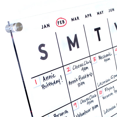 The Clear Acrylic Monthly Wall Calendar by Girl Friday Shop showcases a February layout with highlighted entries like Annie Birthday! on the 1st, and events such as Chess Club and Field Trip. Designed for wet-erase markers, it features columns from Sunday to Saturday and February is circled in red at the top.