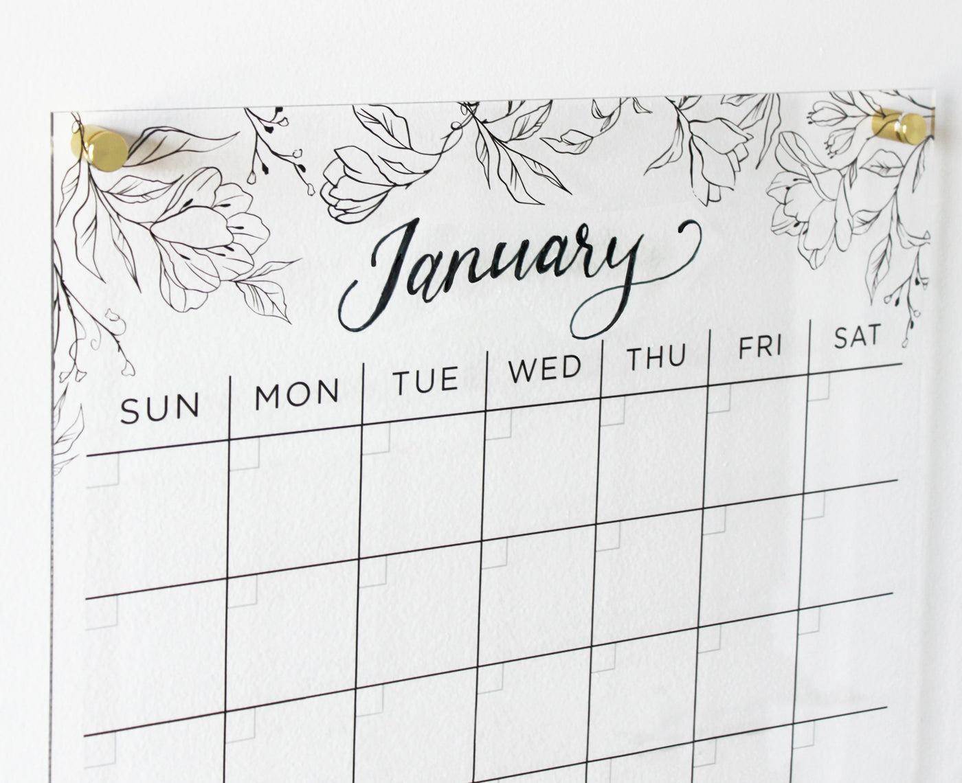 A minimalist Clear Acrylic Monthly Wall Calendar by Girl Friday Shop, showcasing an elegant January scripted over a floral print. Days of the week appear without dates or events, pinned to a white wall with premium hardware.