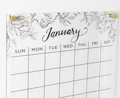 A minimalist Clear Acrylic Monthly Wall Calendar by Girl Friday Shop, showcasing an elegant January scripted over a floral print. Days of the week appear without dates or events, pinned to a white wall with premium hardware.