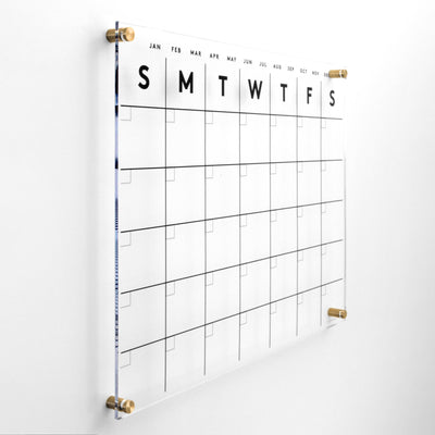 Girl Friday Shops Clear Acrylic Monthly Wall Calendar (Horizontal) has weekday columns and five-week rows. It features durable gold standoff mounts, black month names at the top, and is compatible with wet-erase markers for versatile planning.