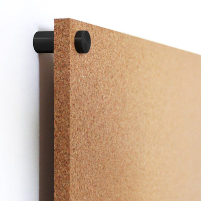A close-up of the Modern Cork Board with dots by Girl Friday Shop shows its textured cork corner mounted on a wall. A black circular bracket holds it in place, allowing for personalization with push pins, against a backdrop where the wall subtly peeks through.
