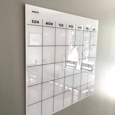 The Girl Friday Shops White Acrylic Monthly Calendar features a wall-mounted dry erase surface with a blank grid titled MONTH and days of the week from Sunday to Saturday. Ideal for meal coordination, it perfectly complements non-magnetic fridges, dining tables, and chairs.