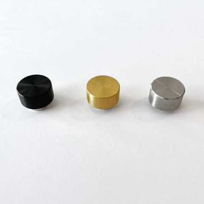 Three smooth, reflective cylindrical knobs, appearing like neodymium magnets, are lined up horizontally on a white acrylic fridge calendar from Girl Friday Shop. They are black, gold, and silver from left to right.