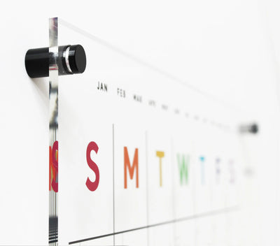 Close-up of a Girl Friday Shops Clear Acrylic Monthly Wall Calendar with 2 Sections, featuring days S, M, T, and W in vibrant red, orange, green, blue. Held by sleek black metal standoffs. Ideal for wet-erase markers.