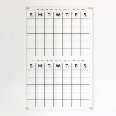 The Clear Acrylic Monthly Wall Calendar by Girl Friday, featuring polished edges and two sections, displays weekdays (S, M, T, W, T, F, S) with months from January to December listed above. It is crafted for a clean and modern design with high-quality materials.