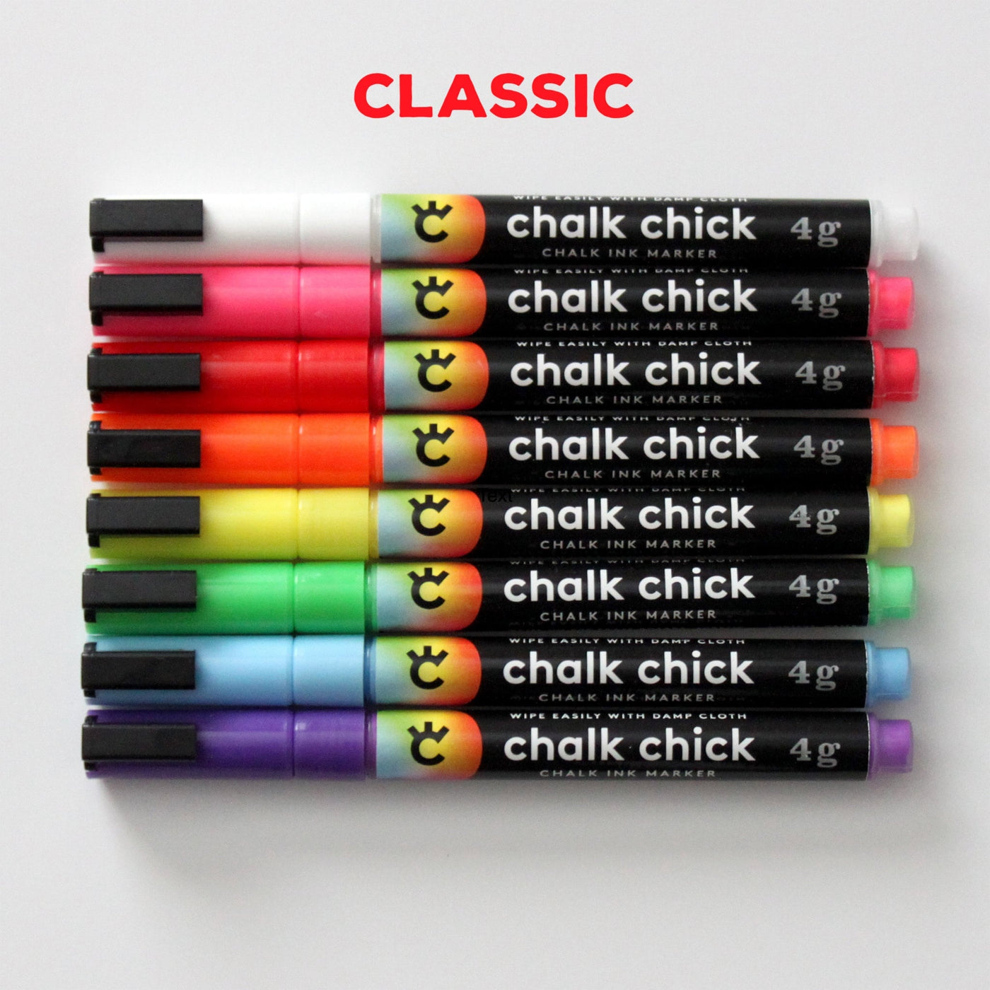 An elegant row of eight 4g Chalk Chick fine tip markers, including vibrant shades like white, pink, and more, are showcased in a sleek Girl Friday Stainless Steel Marker holder. CLASSIC is boldly written above in red.