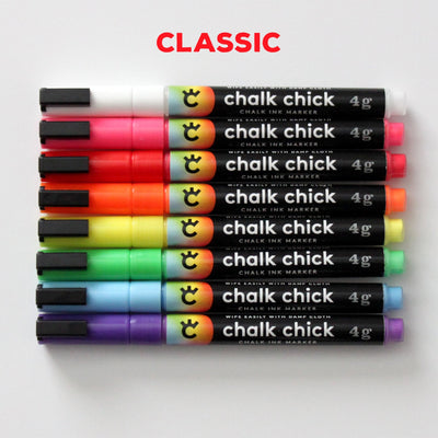 An elegant row of eight 4g Chalk Chick fine tip markers, including vibrant shades like white, pink, and more, are showcased in a sleek Girl Friday Stainless Steel Marker holder. CLASSIC is boldly written above in red.