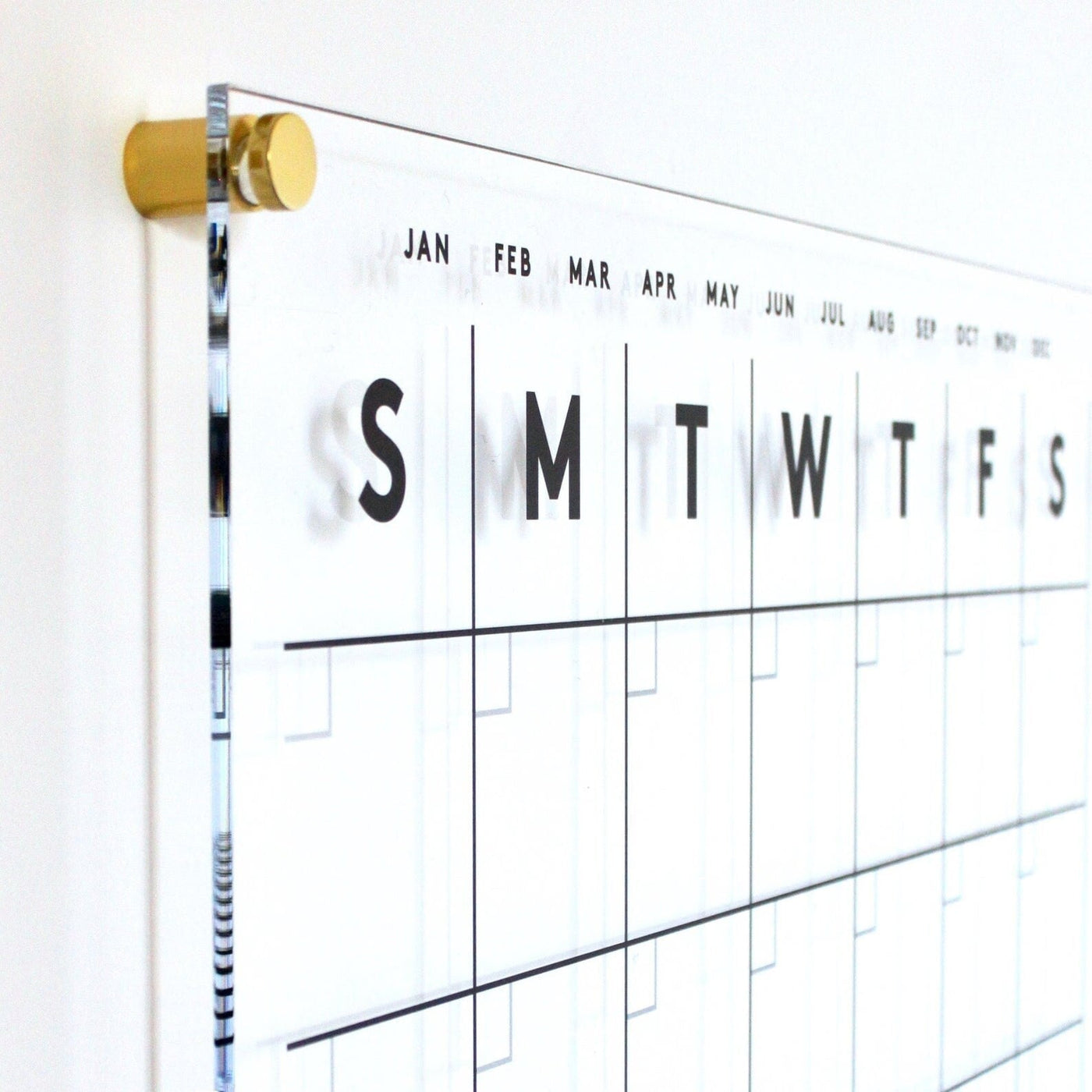 The Girl Friday Clear Acrylic Customizable Monthly & Weekly Wall Calendar, featuring abbreviated weekday names starting with Sunday and months along the side, mounts with gold fixtures on a white wall. Perfect for wet-erase markers to effortlessly update your plans.