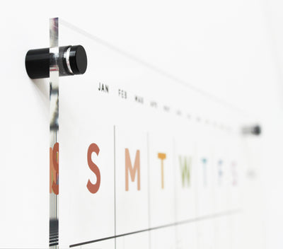 A close-up of the Girl Friday Clear Acrylic Monthly Wall Calendar, Earth Tone Rainbow edition, shows black mount fixtures. It features colorful large letters for days starting with S for Sunday and months in smaller black font from January to May.