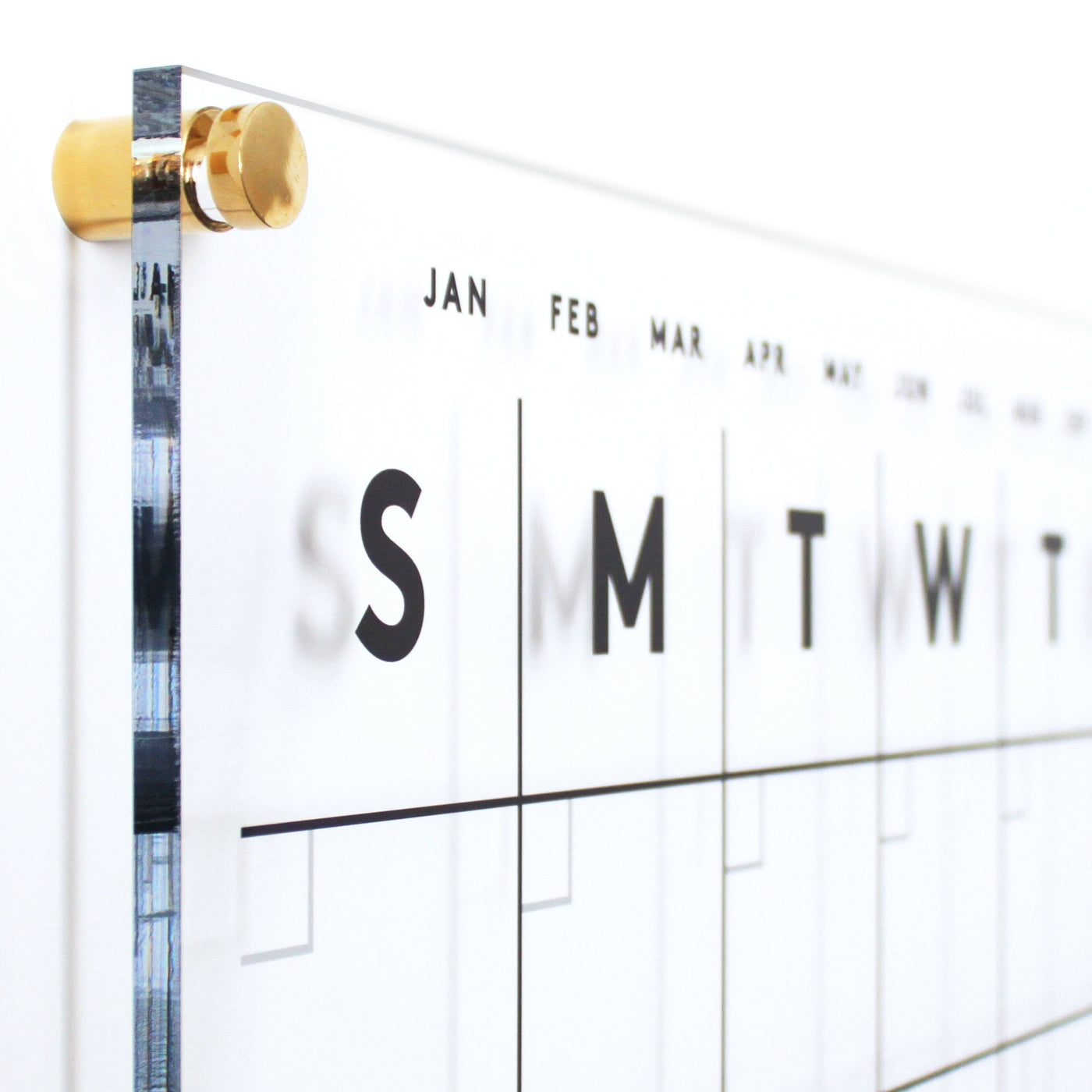 The Girl Friday Clear Acrylic Monthly Wall Calendar features a sleek, minimalist design with polished edges and gold hardware, showcasing days and months in large bold letters. Its modern look against a white background highlights high-quality materials, covering two months at once.